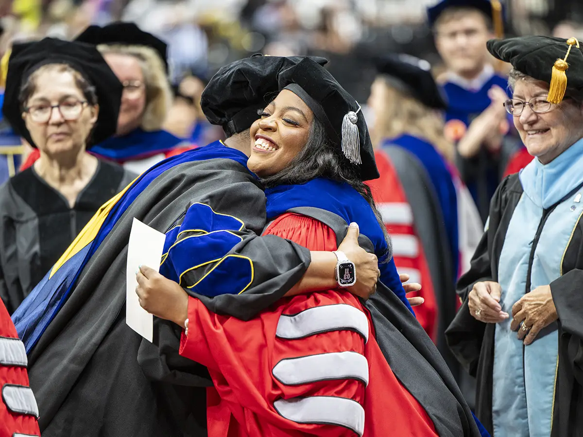 A graduating college student wearing a cap and gown hugs a faculty member at commencement