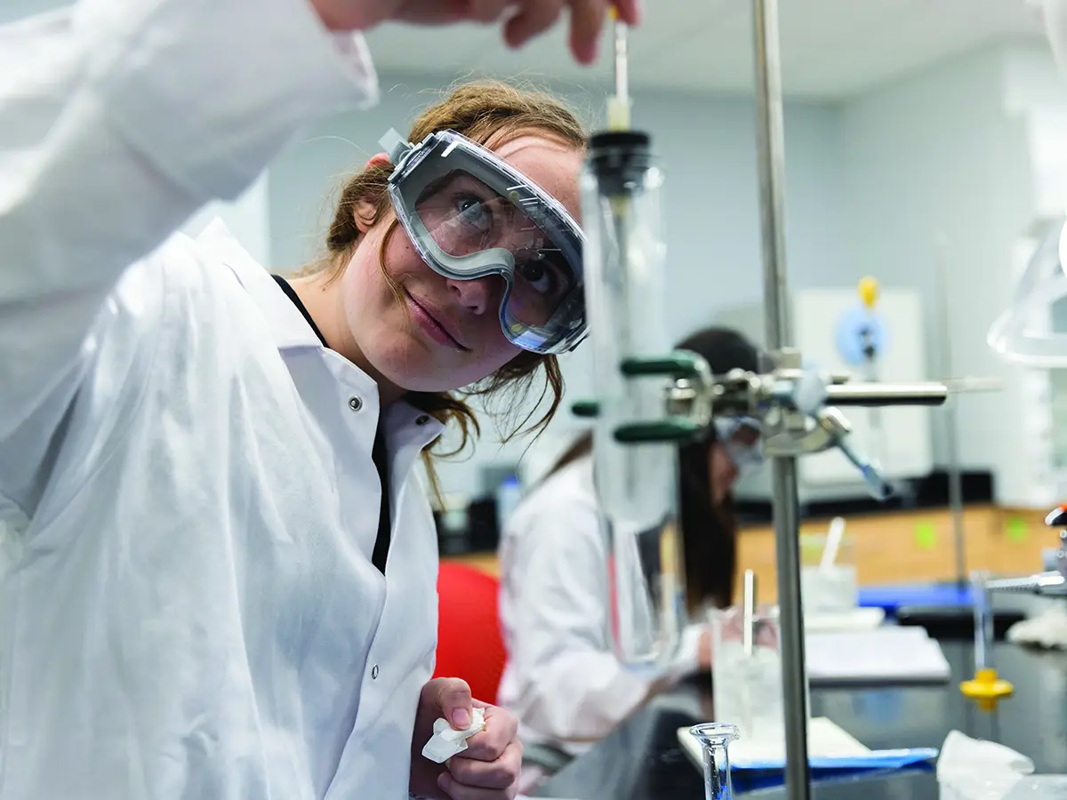 A college student wearing a lab coat and safety goggles manipulates a test tube clamped in a stand in a research lab.
